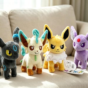 May include: Four plush toys of various Pok&eacute;mon characters are displayed on a beige couch. The plushies include a black Umbreon, a cream-colored Leafeon, a yellow Jolteon, and a purple Espeon. Each has distinctive features and colors.