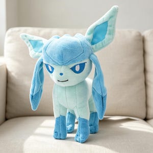 May include: A plush Glaceon toy in shades of blue and light blue, standing on a beige surface. The plush features large ears, a pointed head, and a friendly expression. The toy is a character from the popular Pokemon franchise.