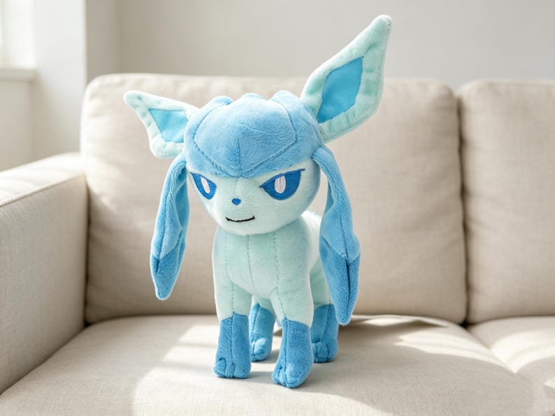 May include: A plush Glaceon toy in shades of blue and light blue, standing on a beige surface. The plush features large ears, a pointed head, and a friendly expression. The toy is a character from the popular Pokemon franchise.