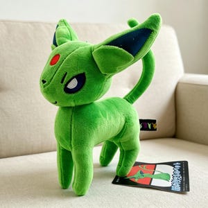 May include: A green plush toy of a creature with large ears, a curved tail, and a red spot on its forehead. The toy has a tag with Japanese text and is on a beige couch.