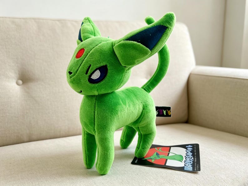 May include: A green plush toy of a creature with large ears, a curved tail, and a red spot on its forehead. The toy has a tag with Japanese text and is on a beige couch.