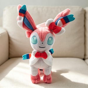 May include: A plush Sylveon toy, featuring a pink, white, and blue color scheme. The toy has large, expressive teal eyes, a red bow, and striped accents on its ears and appendages. It is standing on a beige couch.