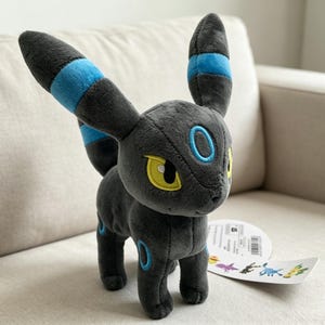 May include: A plush toy of a black creature with large ears and blue accents. The toy has yellow eyes and blue rings on its body. It is sitting on a light-colored couch. A tag with colorful illustrations is attached to the toy.