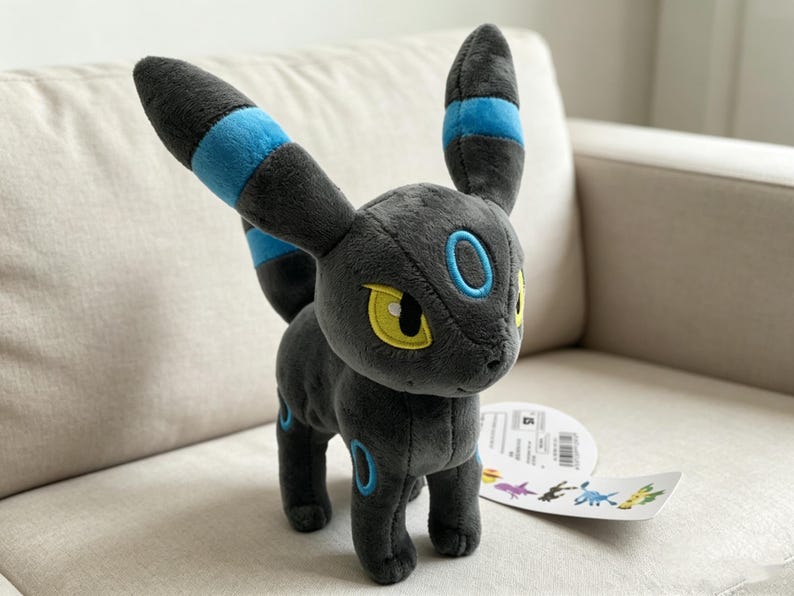 May include: A plush toy of a black creature with large ears and blue accents. The toy has yellow eyes and blue rings on its body. It is sitting on a light-colored couch. A tag with colorful illustrations is attached to the toy.