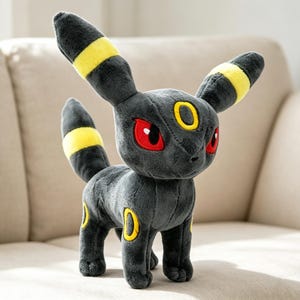 May include: A plush Umbreon toy, featuring a dark gray body with yellow accents on its ears, tail, and circular markings. The toy has red eyes and is standing on a neutral-colored surface.