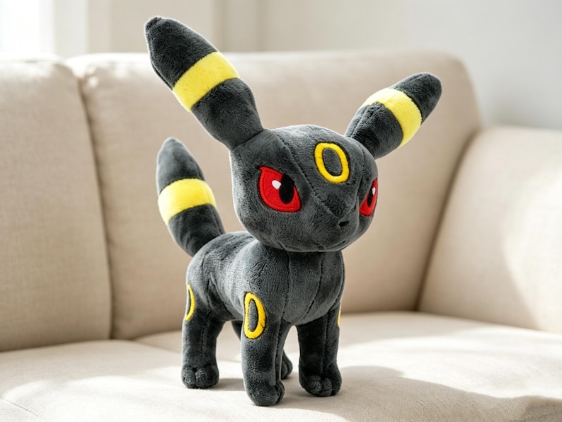 May include: A plush Umbreon toy, featuring a dark gray body with yellow accents on its ears, tail, and circular markings. The toy has red eyes and is standing on a neutral-colored surface.
