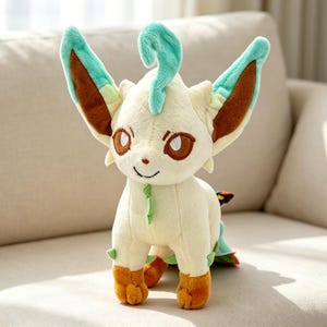 May include: A plush toy of a character with cream-colored fur, brown eyes, and orange feet. The character has large ears with brown and teal accents, and a teal curl on its head. It is sitting on a beige couch.