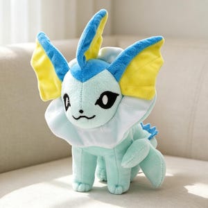 May include: A plush toy of a Vaporeon character, featuring a light blue body, white collar, and blue and yellow fins. The toy has black eyes and a small, embroidered smile. It is sitting on a light-colored sofa.