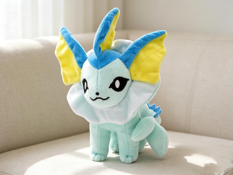 May include: A plush toy of a Vaporeon character, featuring a light blue body, white collar, and blue and yellow fins. The toy has black eyes and a small, embroidered smile. It is sitting on a light-colored sofa.