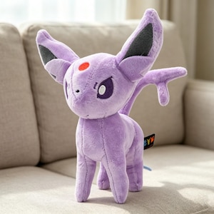 May include: A plush, lavender-colored Espeon toy with large ears, a red spot on its forehead, and a small tail. The toy is standing on a beige couch. The Espeon has purple eyes and a small tag attached to its side.