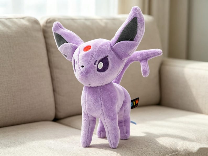 May include: A plush, lavender-colored Espeon toy with large ears, a red spot on its forehead, and a small tail. The toy is standing on a beige couch. The Espeon has purple eyes and a small tag attached to its side.