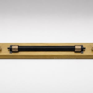 May include: A rectangular brass wall plate featuring a central horizontal black bar held by two ribbed brass cylindrical mounts. Two small brass knobs are positioned on the far left and right sides of the plate, completing the hardware assembly.