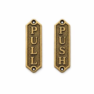 May include: Two rectangular brass-toned metal door plates with pointed ends. One plate features the word PULL in raised lettering, and the other features the word PUSH in raised lettering. Both plates have a textured background and two screw holes.