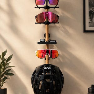 Op de afbeelding: Een verticaal aan de muur gemonteerd houten rek met drie zwarte houders voor sportbrillen, een kleine zwarte plank met een Garmin-apparaat en een zwarte KASK-fietshelm die onderaan de houten constructie hangt.