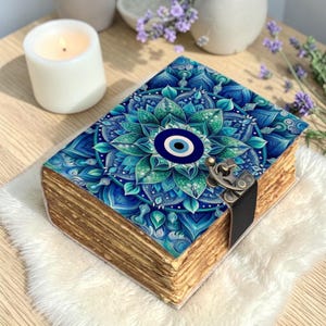 Puede incluir: Un diario decorativo con un diseño de mandala azul y verde que presenta un símbolo de mal de ojo. El diario tiene páginas de aspecto envejecido, un cierre de metal y una correa de cuero negro. Una vela blanca encendida y ramitas de lavanda están en el fondo.