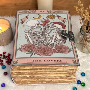 May include: A tarot card with the Lovers design, featuring two skeletons holding flowers, with a light blue background and pink accents. The card is open, revealing the deck's edges. The text "THE LOVERS" is at the bottom.