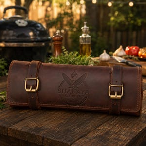 Puede incluir: Un estuche para cuchillos de cuero marrón oscuro con dos correas verticales y hebillas de metal color latón. El centro presenta un logotipo en relieve con dos cuchillos cruzados, una llama y el texto SHANAYA LEATHER HUB HANDMADE.
