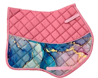 May include: A quilted horse saddle pad featuring a solid pink upper section and a lower section with a blue, purple, and gold marble-like pattern. The pad is edged with pink piping and includes two pink hook-and-loop fastener straps for attachment.