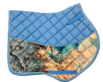May include: A light blue quilted saddle pad featuring a decorative lower panel with a marbled pattern in shades of teal, dark brown, and cream. The pad includes a braided trim along the edge and two blue fabric straps for secure attachment.