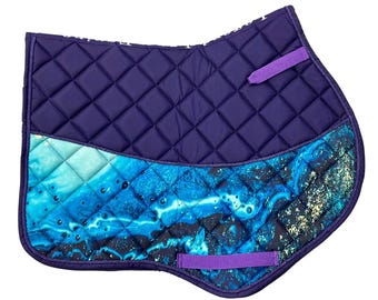 May include: A quilted navy blue saddle pad with a vibrant ocean-themed design. The pad features a diamond pattern, a blue and turquoise wave pattern, and purple accents, including straps for secure placement. The pad is designed for equestrian use.