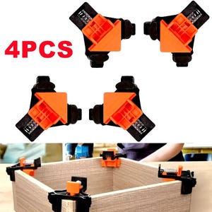 May include: Four black and orange corner clamps with the text "4PCS" in red. These clamps are designed to hold wooden pieces together at a 90-degree angle. The clamps are shown in use on a wooden box.