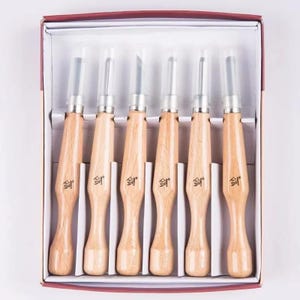 May include: A set of six wood carving chisels with light brown wooden handles and silver metal accents, presented in a white box with a red border. Each chisel has a protective plastic cap.