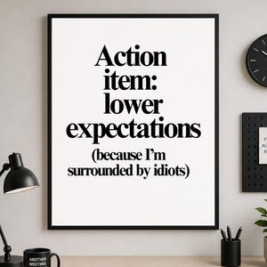 May include: A framed print with the text "Action item: lower expectations (because I'm surrounded by idiots)". A black desk lamp, a black mug, and a black clock are also visible.