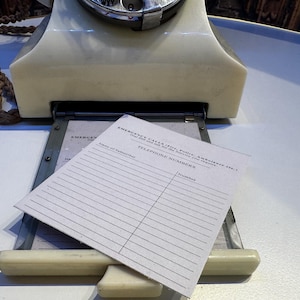 May include: An off-white rotary telephone with a pull-out drawer containing a paper insert. The paper features the text EMERGENCY CALLS, TELEPHONE NUMBERS, Name of Subscriber, and Number. A braided brown cord is attached to the side of the telephone.
