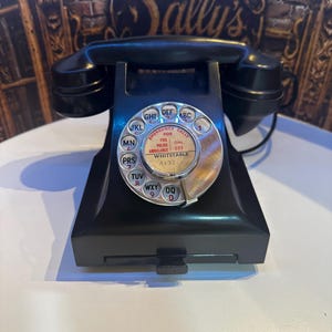 Könnte beinhalten: Ein schwarzes Wählscheibentelefon mit einer kreisförmigen Metallscheibe mit Buchstaben und Zahlen. Die Mitte der Wählscheibe zeigt Text, darunter Emergency Calls, Fire, Police, Ambulance, 999 und Whitstable 4132. Der Hörer liegt auf der Basis.