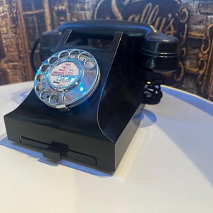 Könnte beinhalten: Ein schwarzes Wählscheibentelefon mit einer Chrom-Wählscheibe und einem zentralen Etikett mit der Aufschrift Emergency Calls For Fire Police Ambulance Dial 999 Southampton. Das Gerät hat einen Hörer oben und ein kleines rechteckiges Schubfach.