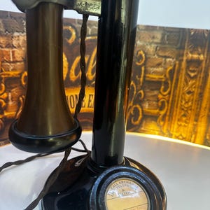 Könnte beinhalten: Ein schwarzes Ständertelefon mit braunem Hörer und geflochtenem Kabel. Der Sockel hat eine runde Anzeige mit dem Text: TO CALL EXCHANGE LIFT RECEIVER & LISTEN, SPEECH ON THIS TELEPHONE IS NEVER SECRET. Die Metallteile sind dunkel gehalten.