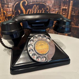Puede incluir: Un teléfono de disco negro vintage con un auricular apoyado en la base. El dial circular incluye números y letras, con una etiqueta central que dice TO CALL EXCHANGE LIFT TELEPHONE & LISTEN y SPEECH ON THIS TELEPHONE IS NEVER SECRET.