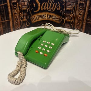 Peut inclure: Un téléphone fixe vert vif avec un combiné assorti et un cordon beige en spirale. L'appareil dispose d'un clavier numérique standard avec des touches blanches et deux boutons orange en bas. Le combiné repose sur l'unité de base.