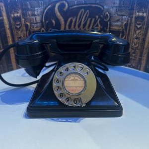 May include: A vintage black rotary telephone featuring a circular silver dial with numbers zero through nine. The centre of the dial includes text that reads TO CALL EXCHANGE LIFT TELEPHONE AND LISTEN, followed by SPEECH ON THIS TELEPHONE IS TO BE KEPT.