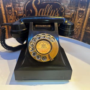 Könnte beinhalten: Ein schwarzes Wählscheibentelefon mit einer goldfarbenen Wählscheibe, die die Zahlen null bis neun zeigt. In der Mitte der Wählscheibe steht der Text: TO CALL EXCHANGE LIFT TELEPHONE, LISTEN, BATH 83019, SPEECH ON THIS TELEPHONE IS NEVER SECURE.