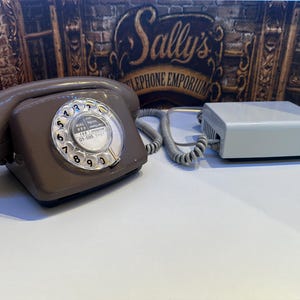 Peut inclure: Un téléphone à cadran taupe avec un disque rotatif numéroté de zéro à neuf et du texte d'urgence. Le téléphone est relié par un cordon torsadé à un boîtier de raccordement rectangulaire gris clair, le tout posé sur une surface blanche.