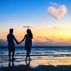 May include: Silhouette of a couple holding hands on a beach at sunset. The sky transitions from blue to orange and yellow, with a heart-shaped cloud above the ocean. The couple is standing in the shallow water, with the waves gently lapping at the shore.