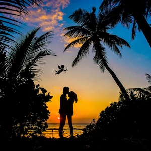 K&ouml;nnte beinhalten: Silhouette eines sich k&uuml;ssenden Paares bei Sonnenuntergang am Strand. Palmen rahmen die Szene ein, mit einem leuchtend orange-gelben Himmel. Eine kleine Silhouette von Amor mit Pfeil und Bogen ist am Himmel.