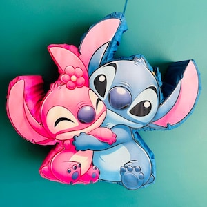 May include: A pi&ntilde;ata depicting two cartoon characters, one pink and one blue, in an embrace. The pink character has a flower on its head. Both characters have large eyes and are smiling. The pi&ntilde;ata is set against a teal background.