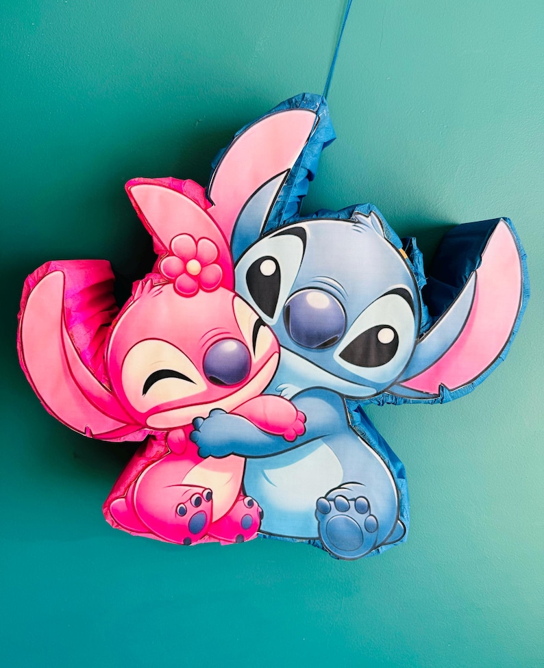 May include: A pi&ntilde;ata depicting two cartoon characters, one pink and one blue, in an embrace. The pink character has a flower on its head. Both characters have large eyes and are smiling. The pi&ntilde;ata is set against a teal background.