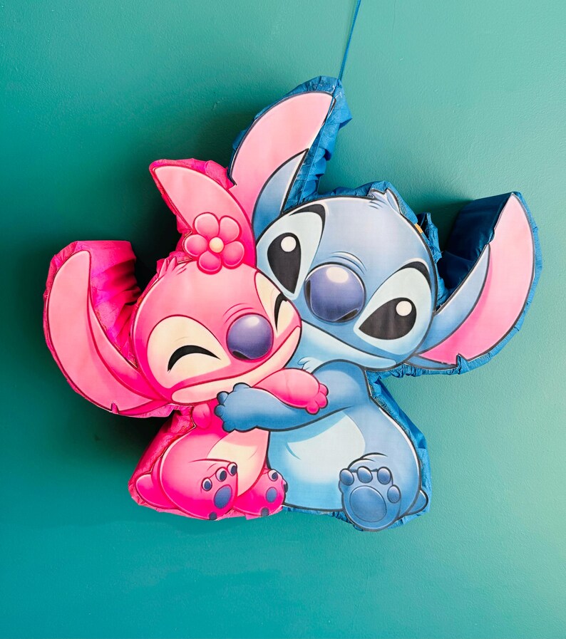 May include: A blue and pink pi&ntilde;ata depicting the characters Stitch and Angel from Lilo & Stitch. Angel, in pink, embraces Stitch, who is blue. The pi&ntilde;ata is set against a teal backdrop.