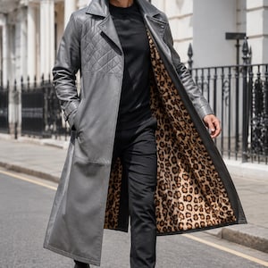 May include: A long grey leather trench coat featuring a quilted pattern on the upper chest and shoulders, a wide collar, and a bold leopard print lining. The coat is worn over a black long-sleeve shirt, black trousers, and black leather boots.