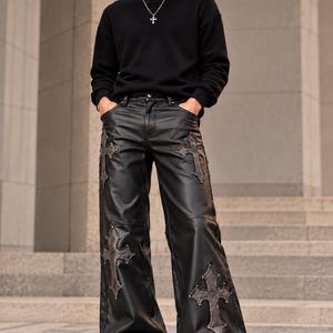 Può includere: Felpa nera a maniche lunghe con girocollo abbinata a pantaloni larghi in ecopelle nera con grandi toppe cucite a forma di croce sulle cosce e sugli stinchi. L'outfit è completato da scarpe da ginnastica bianche e una collana con ciondolo a croce.