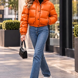 Puede incluir: Una chaqueta acolchada de color naranja brillante con cuello alto, botones de presión frontales y mangas largas, combinada con pantalones vaqueros azules, un jersey de cuello alto negro, zapatillas blancas y un bolso negro pequeño.