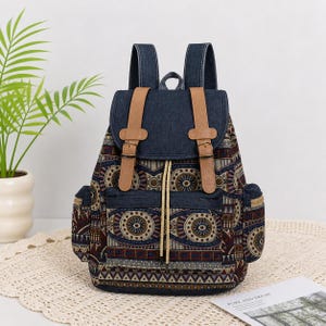 May include: A patterned backpack with a denim flap and straps, featuring brown leather accents and buckles. The bag has a drawstring closure and side pockets. The pattern includes circular and geometric designs in shades of blue, brown, and beige.