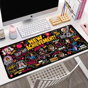 May include: A rectangular black desk mat featuring a vibrant, multi-colored graphic print with the text "NEW ACHIEVEMENT!" and various cartoon illustrations including cats, a dragon, a treasure chest, and phrases like "VIVA LA REVOLUCION CARL".
