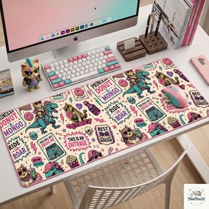 May include: A rectangular desk mat featuring a cream background with a repeating pattern of colorful illustrations including dinosaurs, cats, donuts, skulls, and text such as Princess Donut & Mongo, Ride or Roar, and This Is An Outrage.