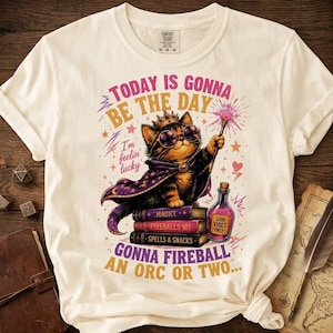 May include: A cream-colored short-sleeve t-shirt featuring a graphic of a cat wearing a crown, sunglasses, and a purple cape. The cat holds a wand over books labeled "MAGIC C", "FIREBALLS 101", and "SPELLS & SNACKS". Text reads "TODAY IS GONNA BE THE DAY I'm feelin' lucky" and "GONNA FIREBALL AN ORC OR TWO..." with a potion bottle.