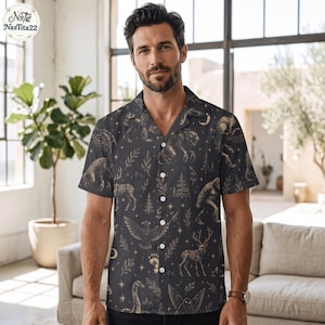 May include: A short-sleeved button-down shirt in dark charcoal gray featuring a repeating gold-toned print of skeletal animals, pine trees, crescent moons, and stars. The shirt has a notched collar and five white buttons down the front center.