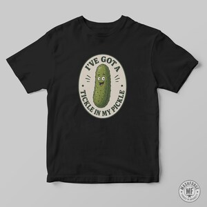 May include: A black short-sleeved t-shirt featuring a central oval graphic with a cartoon green pickle character. The text surrounding the pickle reads I'VE GOT A TICKLE IN MY PICKLE in black capital letters against a light beige background.
